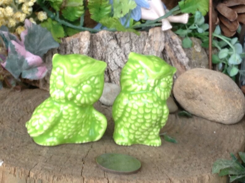 May include: Two green ceramic owl figurines with a textured surface. The owls are sitting on a wooden surface.