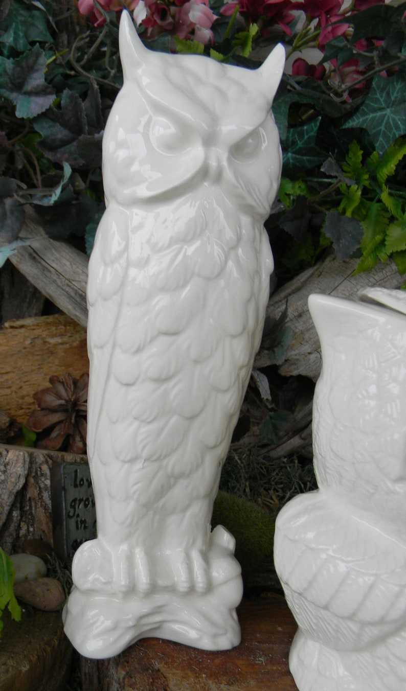 May include: Two white ceramic owl figurines. The owl on the left is standing on a tree branch. The owl on the right is sitting on a tree branch. Both owls have detailed feathers and a realistic appearance.