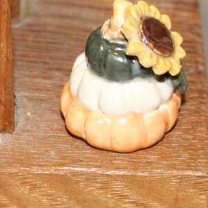 May include: A small ceramic pumpkin figurine with a white pumpkin on top and a green pumpkin on top of that. A yellow sunflower with a brown center sits on top of the green pumpkin.