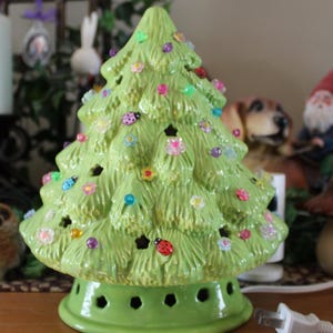 以下が含まれることがあります： 明るい緑色のセラミッククリスマスツリー型のランプ。木にはカラフルな花とてんとう虫が飾られています。ベースには一連の穴があります。白い電気プラグが見えます。