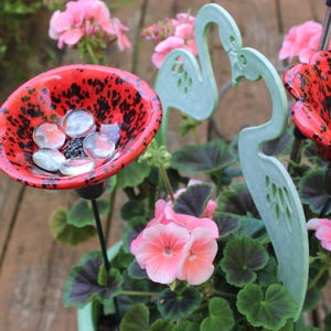May include: A vibrant red and black speckled flower-shaped bird bath filled with clear glass stones. Two light green flamingo garden stakes are positioned near the bird bath, surrounded by pink geraniums and green foliage.