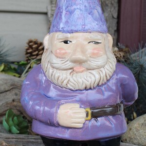 May include: A purple ceramic garden gnome with a white beard and a purple and white hat. The gnome is wearing a purple shirt and brown shoes.
