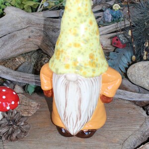May include: A ceramic garden gnome figurine with a yellow hat speckled with orange spots and a long white beard. The gnome wears an orange coat and brown shoes. A small bee charm hangs from the hat.