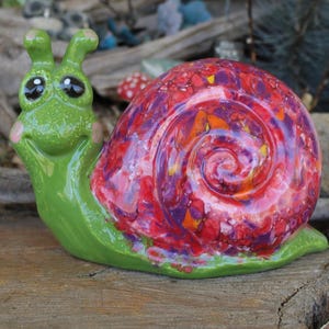 May include: A whimsical ceramic snail figurine with a vibrant, swirled shell in shades of red, purple, and orange. The snail's body is a cheerful green with large, expressive eyes and a smiling face. A decorative garden accent.
