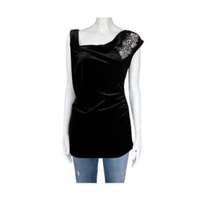 Può includere: Top in velluto nero con scollatura asimmetrica unica e abbellimento decorativo sulla spalla. Il top ha un design drappeggiato ed è esposto su un manichino. La modella indossa jeans blu.