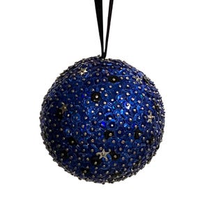 May include: A round, blue Christmas ornament covered in sequins and small star-shaped embellishments. The ornament has a black ribbon for hanging. The sequins are a deep blue color, and the stars are silver.