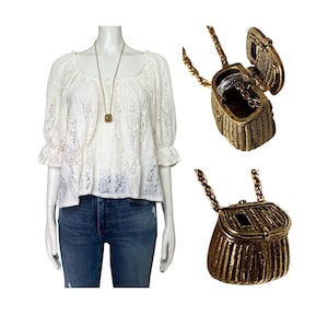 Könnte beinhalten: Eine weiße Spitzenbluse mit quadratischem Ausschnitt und Dreiviertelärmeln wird mit einer blauen Jeans kombiniert. Eine goldfarbene Halskette mit einem kleinen quadratischen Anhänger hängt am Ausschnitt. Eine goldfarbene Handtaschen-Anhänger-Halskette ist ebenfalls abgebildet.