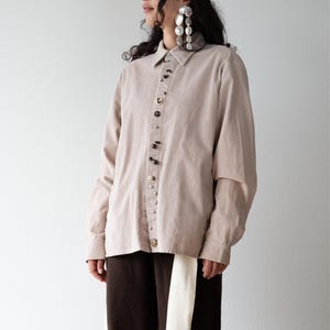 Può includere: Camicia beige di velluto a coste con maniche lunghe e colletto. La camicia è abbottonata con bottoni decorativi. La modella indossa un berretto di maglia marrone scuro e grandi orecchini d'argento. I pantaloni sono marrone scuro con una striscia color crema.