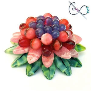 Op de afbeelding: Een handgemaakt bloemenkralenontwerp. De bloem heeft een centrale cluster van paarse en roze kralen, omgeven door lagen roze en groene bloemblad-vormige kralen. Het ontwerp bevat een illustratie van een naald en draad in de rechterbovenhoek.