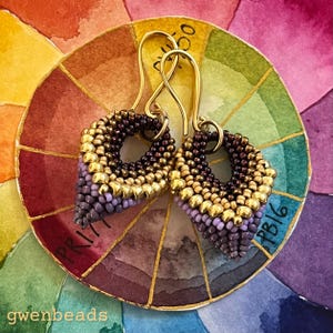 May include: A pair of handmade beaded earrings with gold-colored ear wires. The earrings are a teardrop shape, with a gradient of colors from gold to purple to dark brown. The earrings are displayed on a colorful watercolor color wheel.