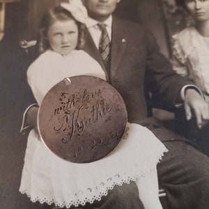 May include: A silver charm with the inscription "with love,  Syathie 12.25.51" hanging from a chain. The charm is in front of a black and white photo of a family.