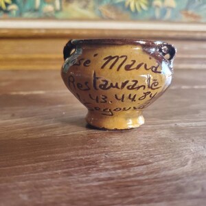 May include: A small, brown ceramic pot with a handle on each side. The pot is decorated with brown lettering that reads "Se. Mend Restaurant 43.448" and "Segourd".