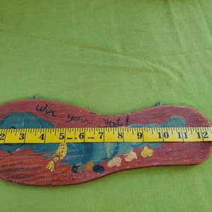 May include: Wooden welcome sign shaped like a foot, painted with a blue and green design and the words "Wipe your feet!". The sign is approximately 33 cm long.