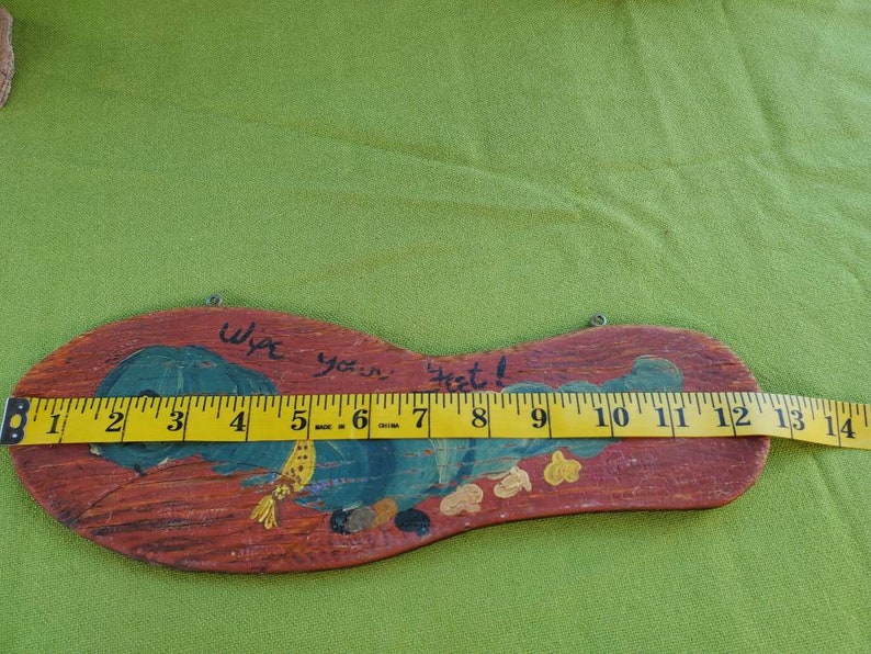 May include: Wooden welcome sign shaped like a foot, painted with a blue and green design and the words "Wipe your feet!". The sign is approximately 33 cm long.