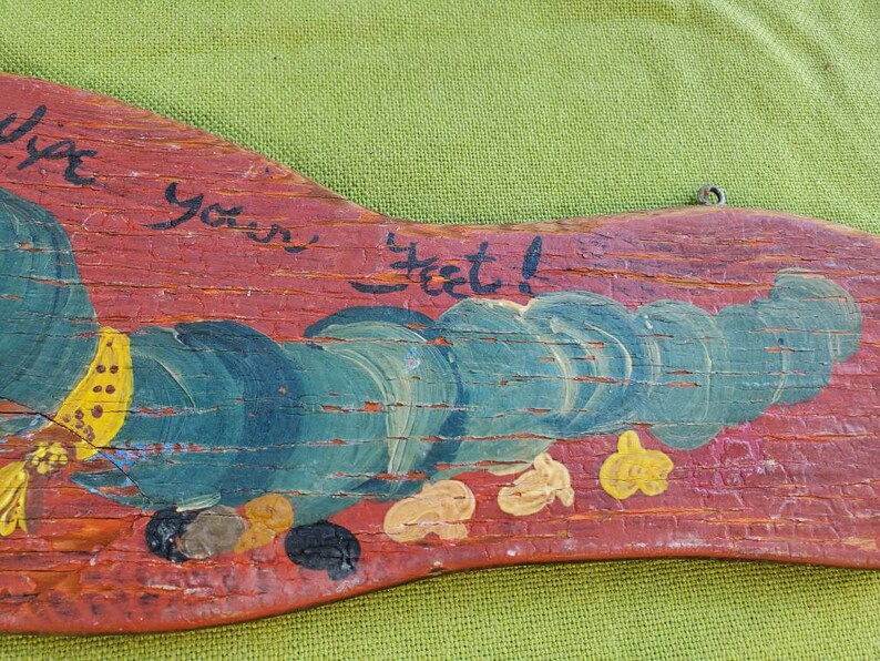 May include: A hand-painted wooden sign with a red background and the words "Wipe your feet!" in black script. The sign features a whimsical design with blue and yellow painted details, and a metal loop for hanging.