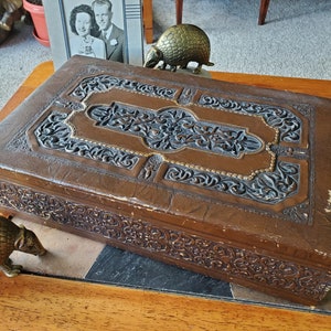 May include: A vintage, brown leather box with intricate embossed floral designs. The box has a gold-colored trim and is in good condition.