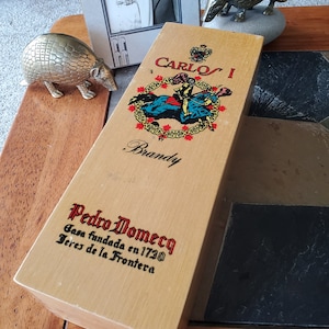 May include: A wooden box with a label that reads "Carlos I Brandy Pedro Domecq Casa fundada en 1730 Jerez de la Frontera". The box is decorated with a colorful image of a knight riding a horse.