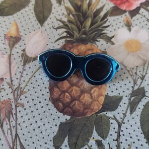 May include: A blue enamel pin shaped like a pair of sunglasses with black lenses. The pin is sitting on a floral patterned fabric with a pineapple in the background.