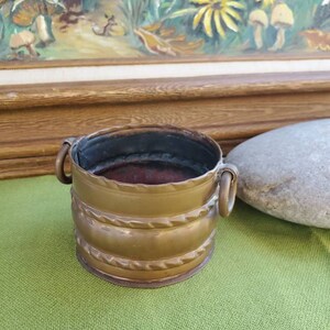 May include: A small, brass planter with two handles. The planter has a textured, rope-like design around the middle.