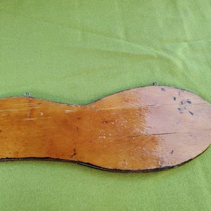 May include: A wooden sign shaped like a shoe sole, with a light brown finish and visible wood grain. It has a metal wire for hanging and shows signs of wear and tear.