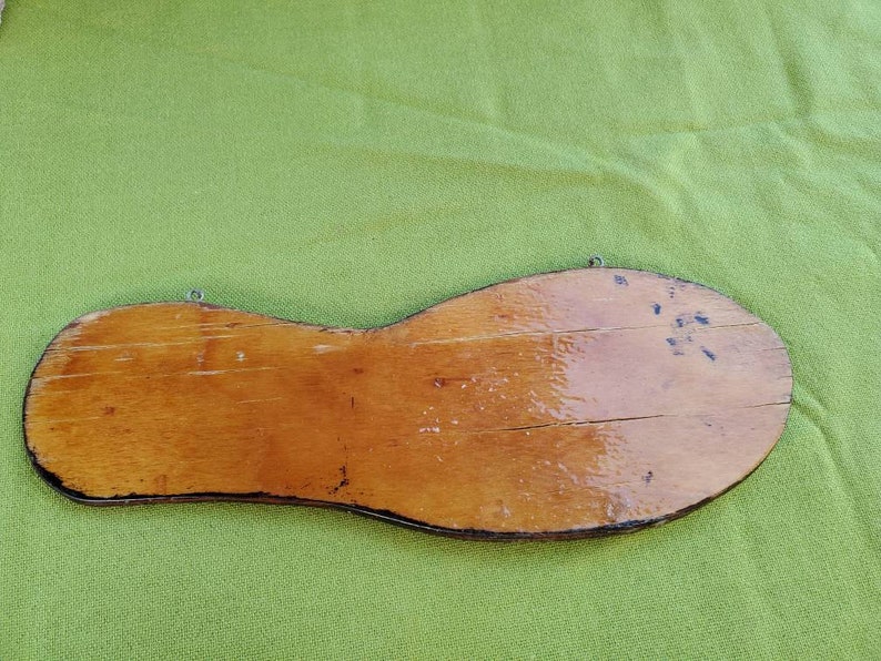 May include: A wooden sign shaped like a shoe sole, with a light brown finish and visible wood grain. It has a metal wire for hanging and shows signs of wear and tear.