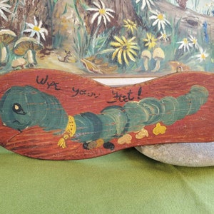 May include: Wooden foot-shaped sign featuring a painted green caterpillar and the words "Wipe your feet!". The sign is painted on a reddish-brown background and has a metal loop for hanging. A charming home decor item.