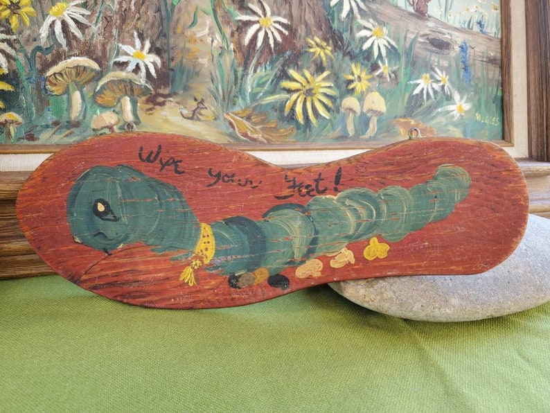 May include: Wooden foot-shaped sign featuring a painted green caterpillar and the words "Wipe your feet!". The sign is painted on a reddish-brown background and has a metal loop for hanging. A charming home decor item.