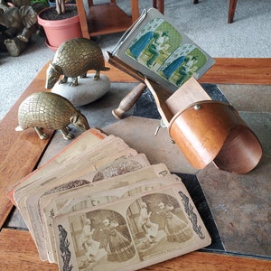 May include: A vintage stereoscope viewer with a wooden frame and a brown leather lens cover. The viewer is holding a stereoscopic card with a black and white image of two people walking down a street. The card has the text "An Unpardonable Mistake" printed on it. There are several other stereoscopic cards stacked on a wooden table next to the viewer. Two brass armadillo figurines are also on the table.
