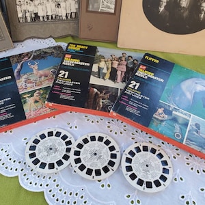 Puede incluir: Tres carretes View-Master vintage con imágenes coloridas. Los carretes están etiquetados como "Bambi", "The Brady Bunch" y "Flipper". Cada carrete tiene el número "21" impreso, que indica el número de imágenes en el carrete. Los carretes están sobre un mantel de encaje blanco.