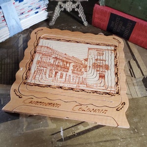 May include: A wooden, laser-cut, framed picture of a building with the text "Chateau Colonar" and "Place St. Francois" on the bottom. The picture is in a light brown frame with a wavy edge.