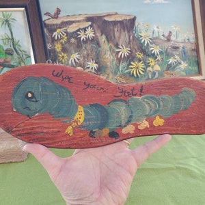 May include: A wooden sign in the shape of a sandal, hand-painted with a green caterpillar and the words "Wipe your feet!" in black. The sign is set against a reddish-brown background, with a yellow collar.