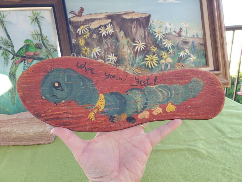 May include: A wooden sign in the shape of a sandal, hand-painted with a green caterpillar and the words "Wipe your feet!" in black. The sign is set against a reddish-brown background, with a yellow collar.