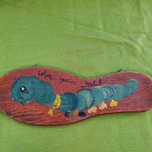May include: Wooden sign shaped like a shoe sole, featuring a blue caterpillar design and the text "Wipe your feet!". The caterpillar has a yellow scarf, set against a red-brown background.