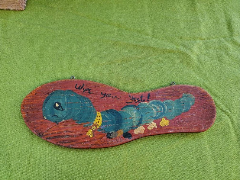 May include: Wooden sign shaped like a shoe sole, featuring a blue caterpillar design and the text "Wipe your feet!". The caterpillar has a yellow scarf, set against a red-brown background.