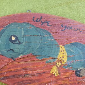 May include: A distressed wooden plaque featuring a painted blue caterpillar with a yellow collar. The caterpillar is set against a red background. The text "Wipe your..." is hand-painted above the caterpillar.