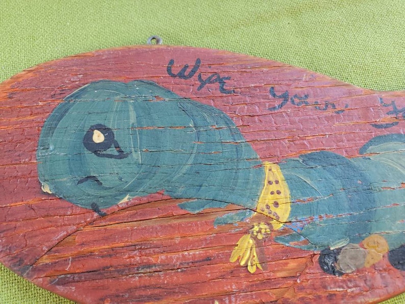 May include: A distressed wooden plaque featuring a painted blue caterpillar with a yellow collar. The caterpillar is set against a red background. The text "Wipe your..." is hand-painted above the caterpillar.