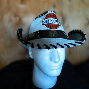 May include: White cowboy hat with black stripes and a brown band. The hat has a red, white, and blue patch that says "Genuine Shit Kicker Made in USA".