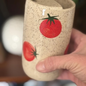 May include: A speckled beige ceramic utensil holder with red tomato designs. The holder has a curved shape with a hole at the top. The tomatoes have green stems and leaves. The holder is being held by a person.