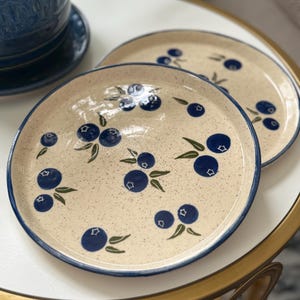 May include: Two ceramic plates with a speckled beige surface and a blue rim. The plates are decorated with a pattern of dark blue blueberries and green leaves. The plates are on a white and gold table.