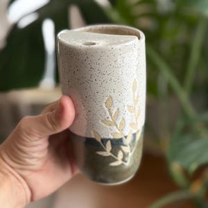 May include: A ceramic tumbler with a speckled, off-white upper half and a dark green lower half. The tumbler features a leaf design in a light tan color. The tumbler is held in a person's hand.