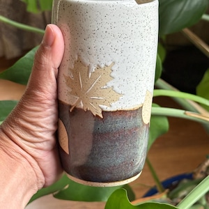 May include: A cylindrical ceramic travel mug featuring a two-tone glaze. The upper portion is white with dark speckles, while the lower section is a deep, earthy brown and green blend. Embossed maple leaf motifs decorate the exterior surface.