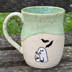 May include: A ceramic mug with a speckled cream-colored base and a light green glazed top. The front features a hand-painted white ghost holding an orange flower, accompanied by two black bats flying above it. The mug has a curved handle on the left.