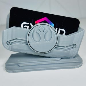 May include: A light grey 3D-printed card holder featuring a circular emblem and the word "GYND" on a black card. The holder has a geometric design and is angled for display.