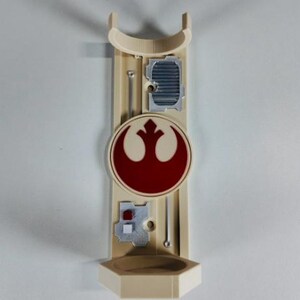 May include: A beige wall-mounted lightsaber holder featuring a red and white Rebel Alliance symbol. The holder has a curved top, a small shelf at the bottom, and several silver-colored details. The design is inspired by the Star Wars franchise.