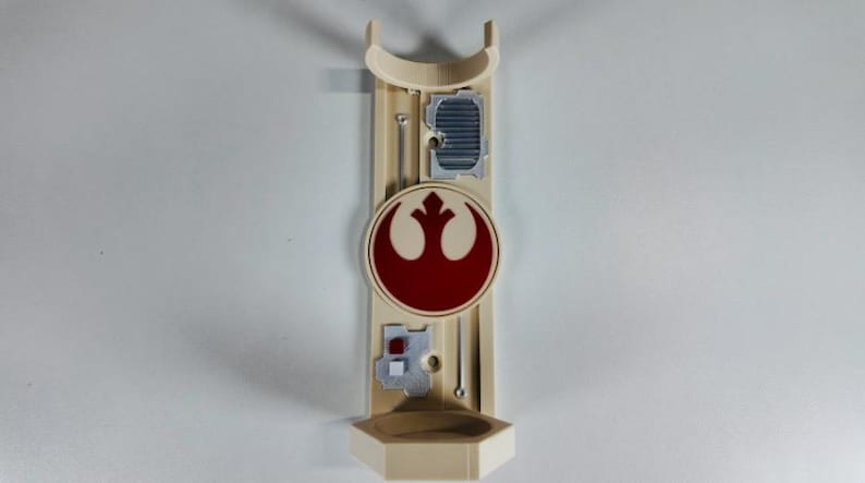 May include: A beige wall-mounted lightsaber holder featuring a red and white Rebel Alliance symbol. The holder has a curved top, a small shelf at the bottom, and several silver-colored details. The design is inspired by the Star Wars franchise.