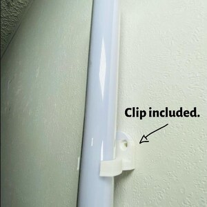 May include: A white, cylindrical light fixture is mounted on a textured wall with a white clip. The text "Clip included" is next to the clip. The fixture is a long, vertical tube.