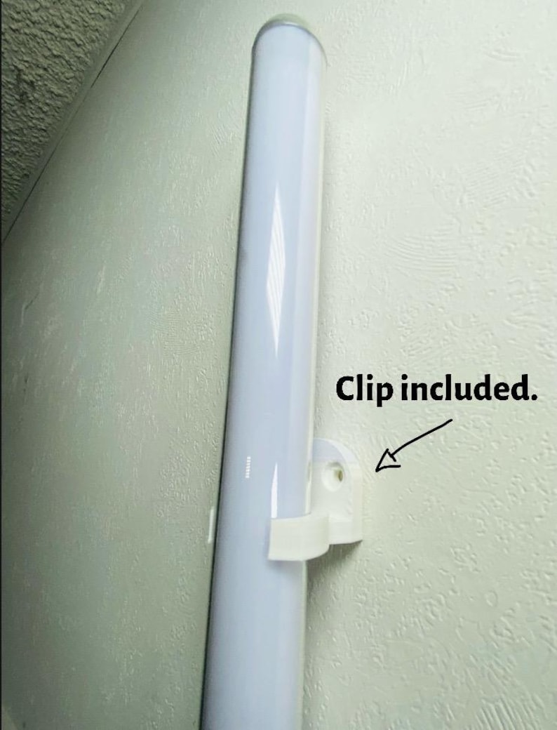 May include: A white, cylindrical light fixture is mounted on a textured wall with a white clip. The text "Clip included" is next to the clip. The fixture is a long, vertical tube.