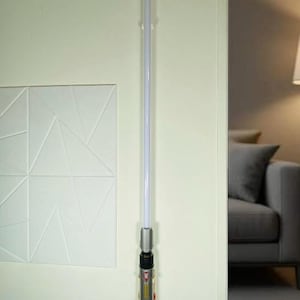 May include: A lightsaber with a silver and black hilt and a white blade is mounted on a wall. The hilt has red and yellow accents. The lightsaber is a prop or toy, likely from a science fiction franchise.