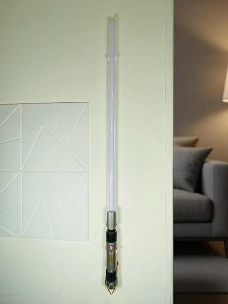 May include: A lightsaber with a silver and black hilt and a white blade is mounted on a wall. The hilt has red and yellow accents. The lightsaber is a prop or toy, likely from a science fiction franchise.