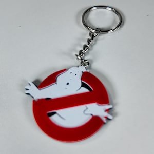 May include: A Ghostbusters keychain featuring the iconic logo. The keychain has a red circle with a white ghost figure and a red slash through it. The keychain is attached to a silver chain and ring.